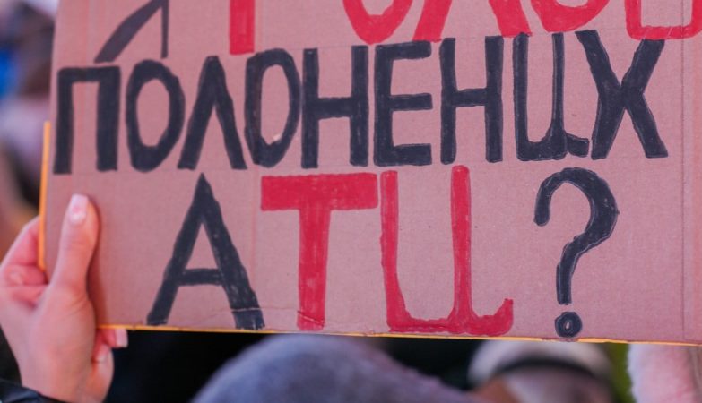 Close-up of a hand holding a handmade cardboard protest sign lettered in red and black Ukrainian text asking who will be the voice of captives. The crowd behind is softly blurred, with hints of blue and yellow flags at a nighttime anti-war rally. Protesters rally in Times Square, New York City, on February 15, 2022, standing in solidarity with Ukraine and speaking out against the war at an anti-war demonstration. Photo: Olek Buzunov (@buzunov.photoart)